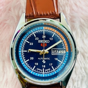 May include: A Seiko 5 automatic watch with a blue dial, silver case, and brown leather strap. The watch face displays the day and date, with the text "SEIKO 5 AUTOMATIC JAPAN MADE". The watch has a silver bezel and a brown leather strap.