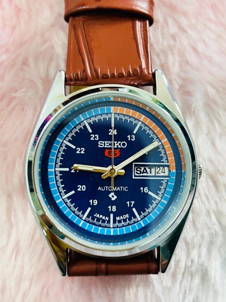 May include: A Seiko 5 automatic watch with a blue dial, silver case, and brown leather strap. The watch face displays the day and date, with the text "SEIKO 5 AUTOMATIC JAPAN MADE". The watch has a silver bezel and a brown leather strap.