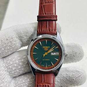 May include: A vintage Seiko 5 automatic watch with a green dial and gold hands. The watch has a date display and is set on a brown leather strap. The watch face is approximately 1.25 inches in diameter.
