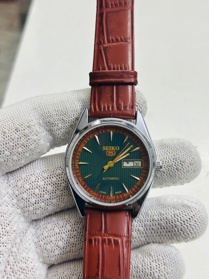 May include: A vintage Seiko 5 automatic watch with a green dial and gold hands. The watch has a date display and is set on a brown leather strap. The watch face is approximately 1.25 inches in diameter.
