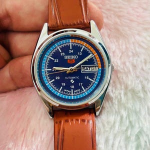 May include: A silver-toned Seiko automatic watch with a blue dial and brown leather strap. The watch face displays the day as "SAT 24" and features white and orange numerals. The watch is a classic timepiece.