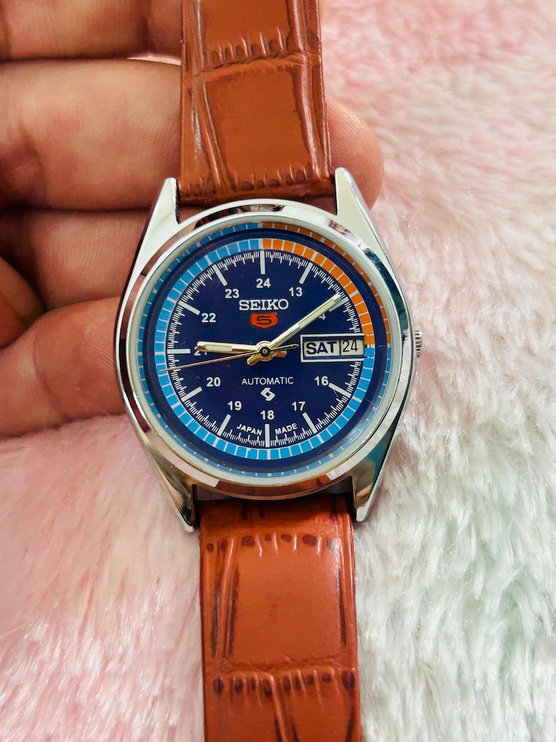 May include: A silver-toned Seiko automatic watch with a blue dial and brown leather strap. The watch face displays the day as "SAT 24" and features white and orange numerals. The watch is a classic timepiece.