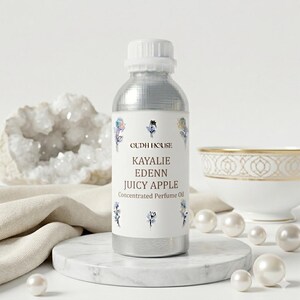 May include: A silver bottle of Kayalie Edenn Juicy Apple Concentrated Perfume Oil. The label features the brand name and product details, with floral accents. The bottle sits on a marble stand, surrounded by pearls and a decorative bowl.