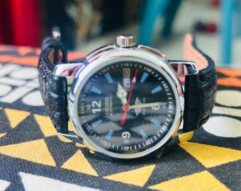 May include: A Citizen automatic wristwatch with a black dial, silver case, and black leather band. The watch face features blue and white markers, a date display, and red second hand. The watch is displayed on a patterned surface.