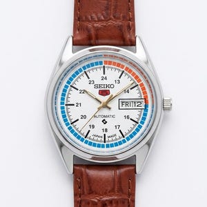 May include: A silver-cased Seiko automatic watch with a white face, blue and orange accents, and a brown leather strap. The watch face displays the day and date, with the text "FRI 12". The watch has a brown leather strap.