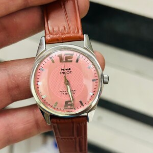 May include: A vintage wristwatch with a silver case and a pink dial. The watch has a brown leather strap and the text "hmt PILOT" on the dial. The watch has a second hand and hour markers.