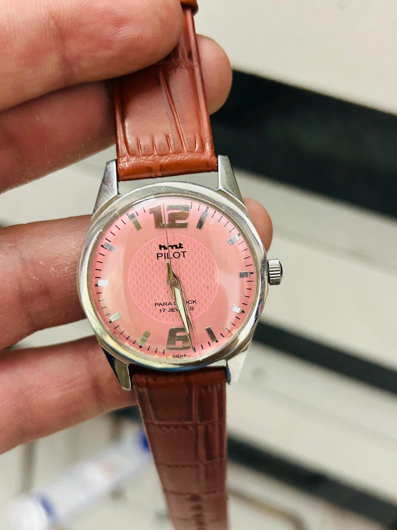 May include: A vintage wristwatch with a silver case and a pink dial. The watch has a brown leather strap and the text "hmt PILOT" on the dial. The watch has a second hand and hour markers.
