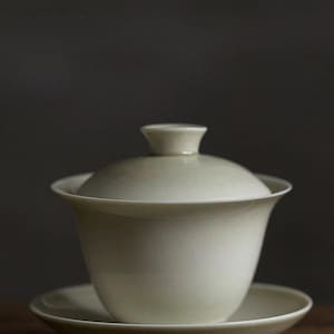 May include: A cream-colored ceramic gaiwan tea set consisting of a bowl, a matching lid with a small knob handle, and a saucer. The tea bowl features a wide, flared rim and a smooth, uniform finish, resting securely on the circular saucer base.