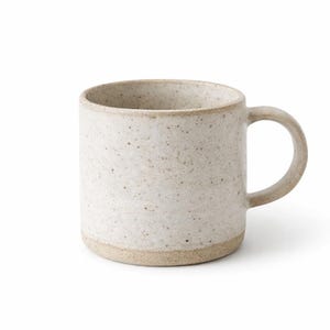 May include: A cylindrical ceramic mug with a speckled off-white glaze and a smooth finish. The base and the curved handle feature an exposed, unglazed tan clay texture, providing a contrast to the speckled body of the drinking vessel.