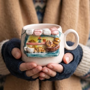 May include: A light pink ceramic mug featuring a three-dimensional sculpted bakery storefront on the front. The miniature shop display includes a teal frame, a striped awning, and tiny clay pastries like croissants and bread loaves on shelves.