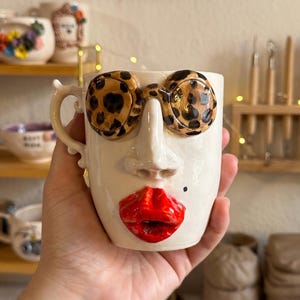 May include: A ceramic mug shaped like a human face with a glossy white finish. The mug features three-dimensional leopard print glasses, a prominent nose, and bright red lips. A small black beauty mark is painted on the right cheek area.