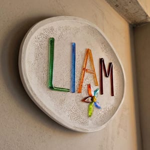 Puede incluir: Una placa ovalada de color blanco roto con textura que muestra el nombre LIAM escrito con varillas de vidrio translúcido de colores. La letra L es verde, I es azul, A es naranja y M es rojo oscuro. Una pequeña flor de vidrio multicolor está bajo la A.