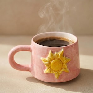 May include: A pink ceramic mug filled with dark coffee, steam rising from the top. The mug features a yellow sun design on the side and a curved handle. The mug is on a neutral background.