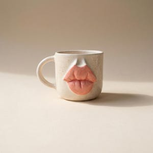 Può includere: Tazza in ceramica maculata con un grande design a labbra rosa in rilievo. La tazza ha un manico bianco e una palette di colori neutri, che la rende un pezzo unico e artistico.