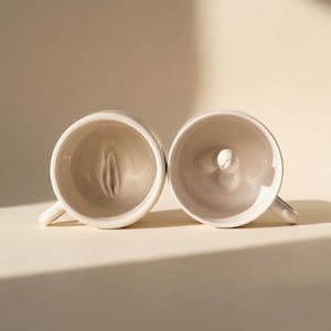 Puede incluir: Dos tazas de cerámica blanca con diseños interiores únicos. Una taza presenta un interior esculpido que se asemeja a una vulva, mientras que la otra tiene un diseño redondeado de tres lóbulos. Ambas tazas tienen asas pequeñas y están sobre un fondo neutro.