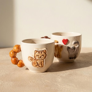 Puede incluir: Dos tazas de cerámica blanquecina. Una taza presenta un diseño de gato, y la otra tiene diseños de corazón y gato. El asa de una taza está hecha de cuentas redondas marrones.