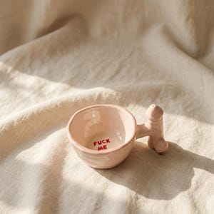 May include: A pale pink ceramic cup with an irregular, hand-formed shape. The interior base features the text FUCK ME printed in red. The handle is sculpted into the distinct shape of a phallus, extending outward from the side of the cup.