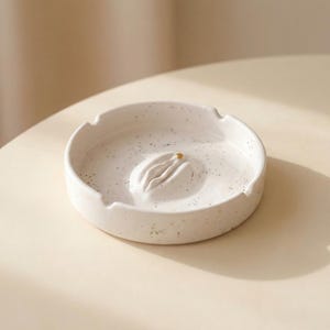 以下が含まれることがあります： 中央に様式化されたデザインと小さな金色のアクセントが付いた、斑点のある白いセラミック灰皿。灰皿は丸い形で、タバコを保持するためのくぼんだ領域があります。