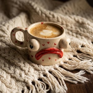 May include: A cream-colored ceramic mug with a handle, featuring two protruding eyes with black heart-shaped pupils and a wide, red, downturned mouth. The mug is decorated with small red hearts and is filled with a coffee beverage with foam.
