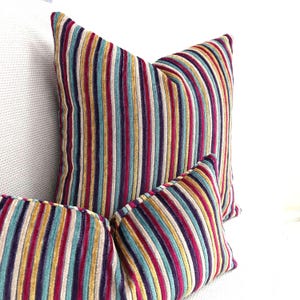 Peut inclure: Un coussin carré et un coussin rectangulaire plus petit présentant un motif à rayures verticales dans des tons de violet profond, magenta vif, jaune moutarde et bleu sarcelle clair. Le tissu a une surface côtelée texturée.
