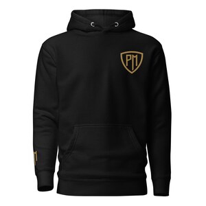Puede incluir: Sudadera con capucha negra con un logotipo dorado en forma de escudo en el pecho y en la manga izquierda. La sudadera tiene un bolsillo delantero y una capucha con cordón.