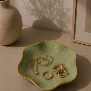 以下が含まれることがあります： 金色の縁取りが施された、波型の淡い緑色のセラミックジュエリー皿。金色のネックレス、指輪、フープイヤリングが置かれている。背景には、ドライフラワーが入ったベージュの花瓶がある。