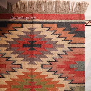 K&ouml;nnte beinhalten: Ein rechteckiger Webteppich mit einem geometrischen Muster in den Farben Rot, Orange, Gr&uuml;n, Schwarz und Beige. Der Teppich hat eine strukturierte Oberfl&auml;che und Fransen an der Oberkante. Der Text indianRugsCraft ist oben links in Wei&szlig; aufgedruckt.