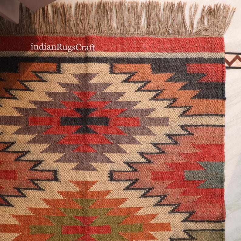 K&ouml;nnte beinhalten: Ein rechteckiger Webteppich mit einem geometrischen Muster in den Farben Rot, Orange, Gr&uuml;n, Schwarz und Beige. Der Teppich hat eine strukturierte Oberfl&auml;che und Fransen an der Oberkante. Der Text indianRugsCraft ist oben links in Wei&szlig; aufgedruckt.
