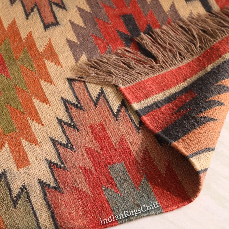 K&ouml;nnte beinhalten: Ein gewebter Teppich mit einem geometrischen Muster aus gezackten, gestuften Formen in Rot-, Orange-, Olivgr&uuml;n- und Dunkelgraut&ouml;nen auf beigem Grund. Der Teppich hat eine umgeschlagene Ecke, die eine strukturierte Unterseite und braune Fransen zeigt.
