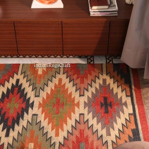 K&ouml;nnte beinhalten: Ein rechteckiger Teppich mit einem sich wiederholenden geometrischen Rautenmuster in Orange-, Rot-, Gr&uuml;n- und Dunkelgraut&ouml;nen auf cremefarbenem Grund. Der Text "indianRugsCraft" ist in Wei&szlig; im oberen Bereich des Teppichs aufgedruckt.