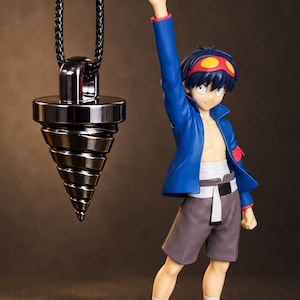 May include: A metallic, gunmetal-colored drill-shaped pendant hangs from a black braided cord. Beside it stands an anime-style figure with dark hair, wearing a blue jacket, grey shorts, and holding a small gold-colored drill accessory.