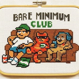 May include: Cross-stitch art in a wooden hoop features a cartoon of a person, cat, and dog on a brown couch. The text "BARE MINIMUM CLUB" is stitched above the couch in black and green.