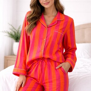 Könnte beinhalten: Ein zweiteiliger Pyjama mit vertikalen Streifen in Orange und Pink. Das langärmlige Oberteil hat eine Knopfleiste mit weißem Paspel, und die Hose hat einen Kordelzug in der Taille. Der Pyjama ist aus einem weichen, bequemen Material gefertigt.