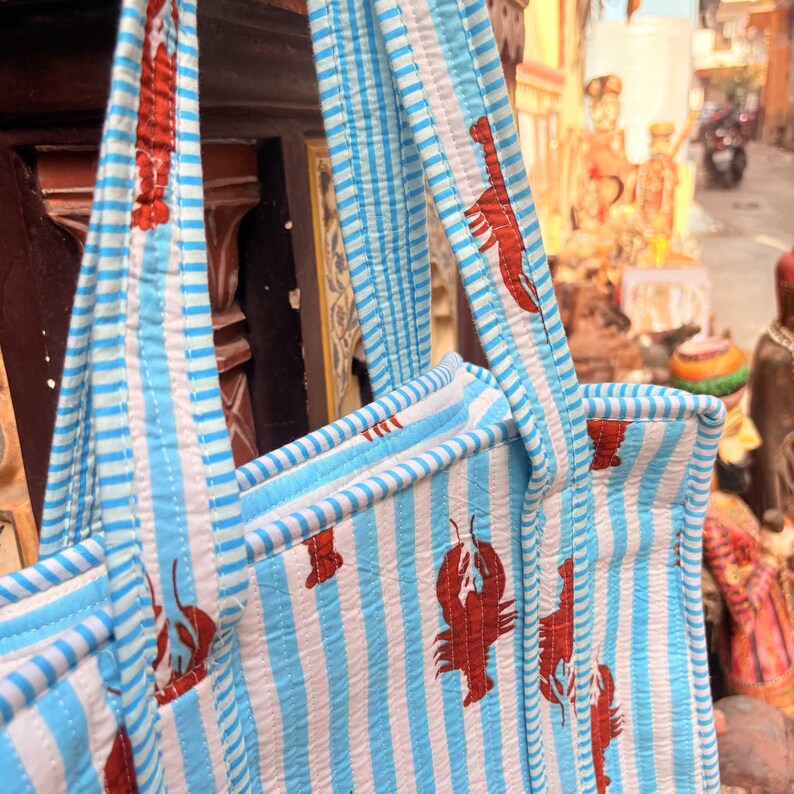 Puede incluir: Primer plano de un bolso tote a rayas azules y blancas con ilustraciones de langostas rojas. El bolso tiene asas a rayas azules y blancas a juego y un dise&ntilde;o acolchado. El bolso est&aacute; hecho de una tela suave y texturizada.