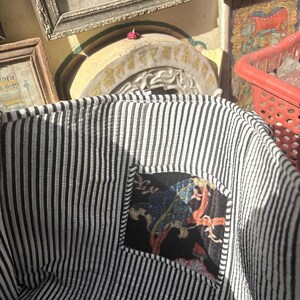 May include: A black and white striped fabric basket with a small, patterned patch. The patch features a colorful design on a black background. The basket is open, revealing the interior lining. A red basket is visible in the background.