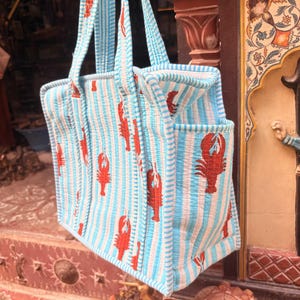 Puede incluir: Un bolso tote a rayas azules y blancas con un estampado de langosta roja. El bolso tiene dos asas y un bolsillo exterior. El bolso est&aacute; hecho de una tela acolchada.