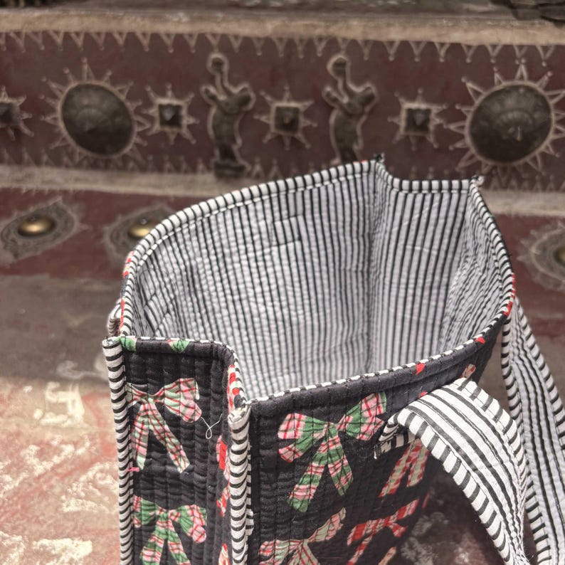 May include: A quilted tote bag featuring a black background with a repeating pattern of red and green plaid bows. The interior and the two shoulder straps are lined with a black and white vertical pinstripe fabric, providing a contrast to the exterior.