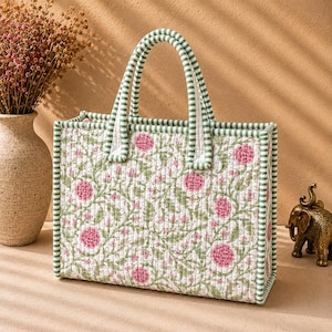 May include: A rectangular quilted tote bag featuring a white base with a repeating pattern of pink flowers and green leafy vines. The bag has two top handles and edges trimmed in a green and white striped fabric, providing a structured shape.