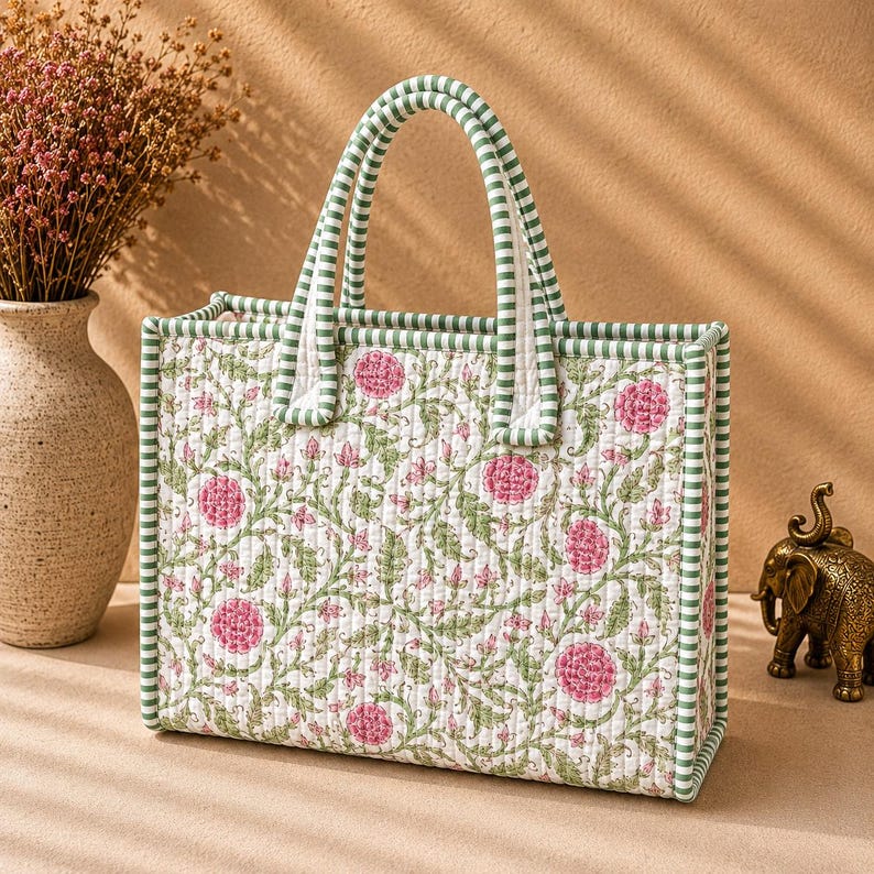 May include: A rectangular quilted tote bag featuring a white base with a repeating pattern of pink flowers and green leafy vines. The bag has two top handles and edges trimmed in a green and white striped fabric, providing a structured shape.