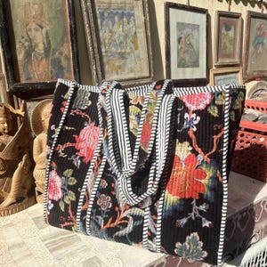 May include: Black tote bag with a floral pattern in red, pink, and blue. The bag has a quilted design with white and black striped accents on the handles and edges. The bag is sitting on a stone surface.