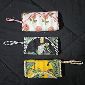 Op de afbeelding: Drie rechthoekige gewatteerde stoffen etuis met polsbandjes en drukknoopsluitingen. Het bovenste etui is wit met roze bloemmotieven en een rood gestreepte rand. Het middelste etui is zwart met een groene apenprint en een zwart-wit gestreepte rand. Het onderste etui is geel met groene palmbomen en een geel gestreepte rand.