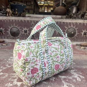 Pode incluir: Uma mala de viagem acolchoada com base branca e um padr&atilde;o floral repetitivo em tons de rosa e verde. A mala inclui duas al&ccedil;as superiores e um fecho de correr, acentuado com debrum de tecido &agrave;s riscas verdes e brancas nas costuras e al&ccedil;as.