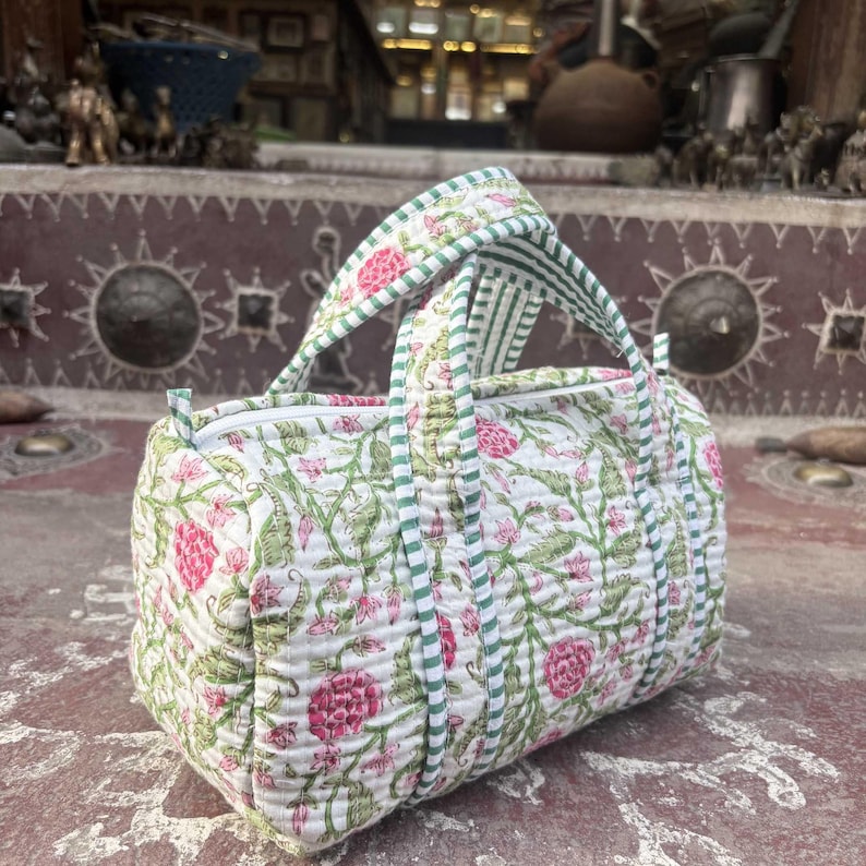 Pode incluir: Uma mala de viagem acolchoada com base branca e um padr&atilde;o floral repetitivo em tons de rosa e verde. A mala inclui duas al&ccedil;as superiores e um fecho de correr, acentuado com debrum de tecido &agrave;s riscas verdes e brancas nas costuras e al&ccedil;as.