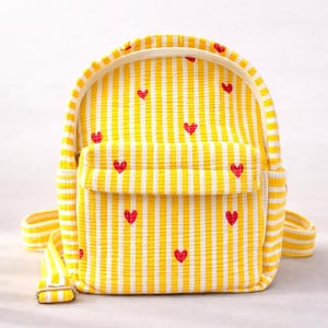 Puede incluir: Mochila a rayas amarillas y blancas con detalles de corazones rojos. La mochila pequeña tiene un bolsillo frontal y correas ajustables. La bolsa está hecha de tela texturizada.