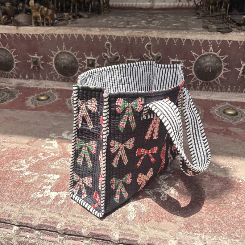May include: A quilted tote bag featuring a black background with a repeating pattern of red, green, and white plaid bows. The bag has black and white striped fabric trim along the edges and matching striped handles, with a striped interior lining.