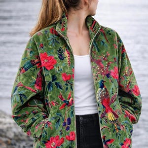 Op de afbeelding: Groene fluwelen jas met ritssluiting en een bloemen- en vogelpatroon. De jas heeft rode bloemen, kleurrijke vogels en donkere bessen op een groene achtergrond. De jas wordt gecombineerd met een zwarte jeans en een witte top.