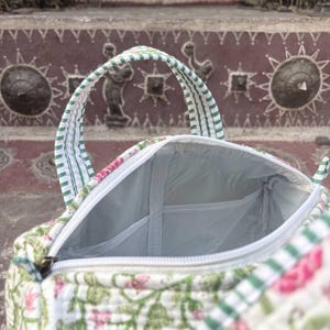 Pode incluir: Uma bolsa de tecido acolchoado com base branca e um padr&atilde;o floral verde e rosa. A bolsa possui duas al&ccedil;as com um design de riscas verdes e brancas, um fecho de correr branco e um forro interior de cor clara com um bolso aberto.