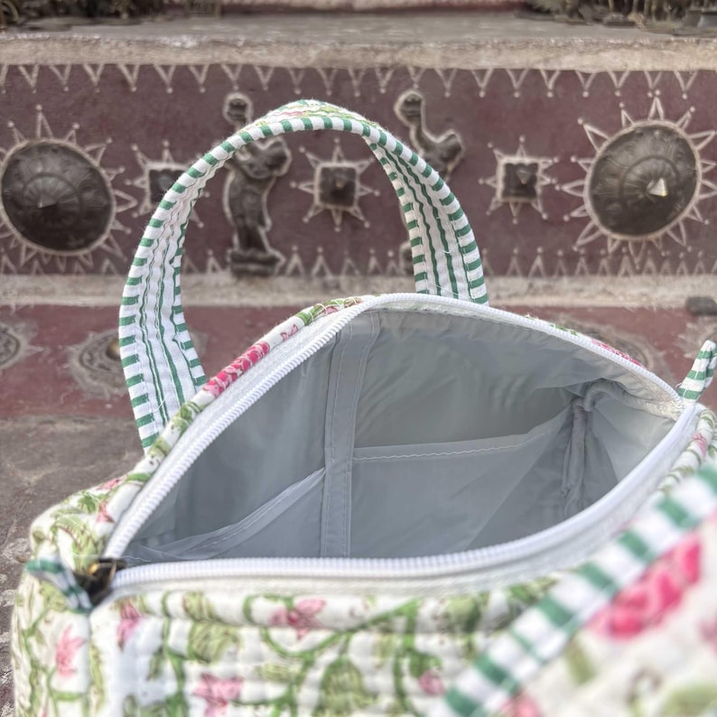 Pode incluir: Uma bolsa de tecido acolchoado com base branca e um padr&atilde;o floral verde e rosa. A bolsa possui duas al&ccedil;as com um design de riscas verdes e brancas, um fecho de correr branco e um forro interior de cor clara com um bolso aberto.