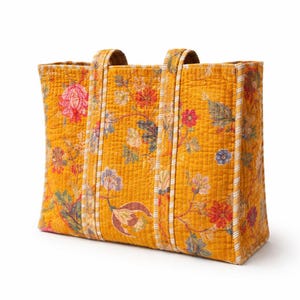 May include: A quilted tote bag in a mustard yellow color, adorned with a floral pattern in shades of pink, red, blue, and green. The bag features two sturdy handles and vertical striped accents. The bag is approximately 15 inches wide, 12 inches tall, and 6 inches deep.