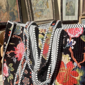 May include: Black tote bag with a floral pattern in red, pink, and green. The bag has a quilted texture and is trimmed with black and white striped fabric. The handles are also striped.